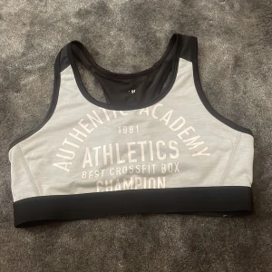 Grå sport-bh med svart kant och tryck - Sport-bh i ljusgrått med svart kant och bred resår.  Racerback-modell för extra stöd, perfekt för träning.