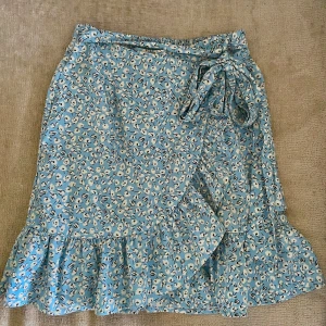 Blå omlottkjol med volang - Söt blå omlottkjol med vitt och svart blommigt mönster. Kjolen har volang längs nederkanten och knyts i midjan. Perfekt för en somrig look och lätt att matcha med en enkel topp🤍🌊