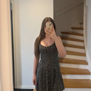 Svart blommig sommarklänning från FB Sister - Supersöt svart klänning med vita små blommor från FB Sister. Klänningen har smala axelband, lätt utställd kjol och en feminin passform. Perfekt för varma dagar och enkel att styla med sneakers eller sandaler. Klänningen har en dragkedja och en gullig design på baksidan. 