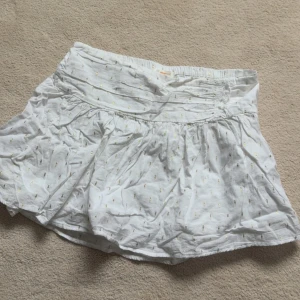 Vit kortkjol med småmönster - Söt vit kortkjol med små diskreta mönster i guld. Kjolen har bred resår i midjan och är lätt utställd för en luftig känsla. Perfekt för varma dagar och enkel att matcha med olika toppar. Använd få gånger, ser väldigt skrynklig ut men de går lätt att stryka!