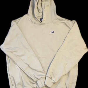 Beige hoodie från New Balance - Säljer en beige hoodie från New Balance med liten logga på bröstet. Tröjan har huva och ribbade muddar vid ärmslut och nederkant. Perfekt för en avslappnad stil och enkel att matcha med det mesta. Priset kan diskuteras och är i PERFEKT skick.