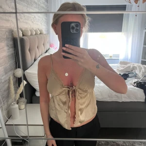 Beige satinlinnetopp med spets och knyt - Säljer en beige linnetopp i satin med tunna axelband, spetsdetaljer och knyt framtill. Toppen har en croppad passform och en lätt glansig yta som ger en lyxig känsla.