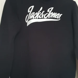 Svart hoodie från Jack & Jones - Svart hoodie från Jack & Jones med vit logga framtill. Klassisk modell med huva och vita dragsnören. Perfekt för en avslappnad stil och enkel att matcha med det mesta. Stolek16år ca xs
