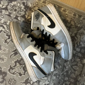 Nike Air Jordans i vitt och grått - Säljer ett par klassiska Nike Air Jordans i vitt och ljusgrått med svarta detaljer och svart swoosh. Skorna har svart snörning, hög modell och rund tå. Perfekta för dig som gillar streetwear och sneakers med ikonisk design.