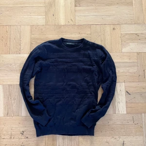 Marinblå stickad tröja från Selected Homme - Säljer en mörkblå stickad tröja från Selected Homme med diskret strukturmönster över bröst och ärmar. Tröjan har rund halsringning och långa ärmar. Perfekt för lager-på-lager under kyligare dagar. 