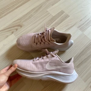 Ljusrosa Nike sneakers - Säljer ett par ljusrosa Nike sneakers med vit sula och klassisk swoosh-logga på sidan. Skorna har snörning och är tillverkade i syntetmaterial med meshstruktur för bra andningsförmåga. Perfekta för dig som gillar stilrena och sportiga skor.