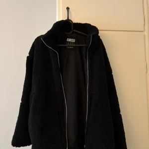 Svart teddyjacka från Amisu - Mysig svart teddyjacka från Amisu med dragkedja framtill. Jackan har två fickor och är tillverkad i ett mjukt, syntetiskt material som ger en fluffig känsla. Perfekt för kyliga dagar. Storlek 38 men passar också 36, har ett litet jack där framme men enkelt o fixa om man vill annars syns de inte
