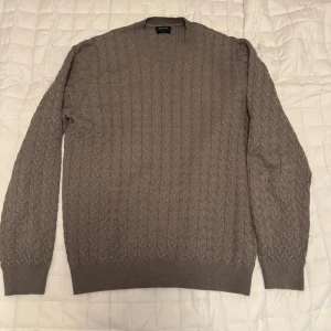 Khaki stickad tröja från Massimo Dutti - Säljer en khaki stickad tröja från Massimo Dutti med ett diskret kabelstickat mönster. Tröjan har rund halsringning och långa ärmar med ribbade muddar. Perfekt för kyligare dagar.