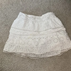 Vit broderad kortkjol med volang - Superfin vit kortkjol med broderade blommor och volangdetaljer. Kjolen har resår i midjan och är gjord i ett lätt bomullstyg, perfekt för varma dagar. Söt och luftig modell som passar till många olika stilar.