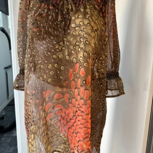 Mönstrad klänning/tunika från Isolde - Säljer en luftig och transparent klänning/tunika från Isolde med unikt fjäderinspirerat mönster i brunt, orange och beige. Klänningen har trekvartsärmar med resår och volangdetalj samt knytning vid halsen. Perfekt för dig som gillar bohemisk stil.