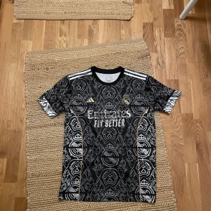 Real Madrid svart bortatröja 2023/24 - Säljer en svart Real Madrid bortatröja från Adidas med unikt mönster i grått och detaljer i guld. Tröjan har korta ärmar, rund hals och tre vita ränder på axlarna. Perfekt för dig som vill sticka ut på läktaren eller planen.