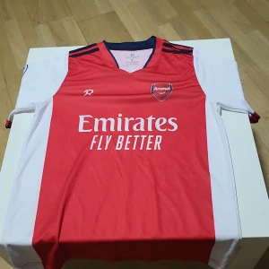 Arsenal hemmatröja Aubameyang 14 - Säljer en Arsenal fotbollströja med Aubameyang 14 på ryggen. Tröjan är röd med vita ärmar och har mörkblå detaljer vid kragen och axlarna. Emirates Fly Better tryck på bröstet och klubbmärke på vänster sida.