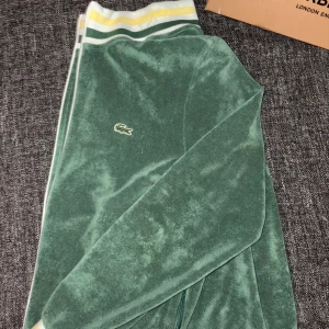 Grön velour tracksuit hoodie/jacka från Lacoste - Grön velourjacka från Lacoste med klassisk krokodillogga på bröstet. Jackan har ribbade muddar med vita, gröna och gula ränder vid kragen och nederkanten. Mjuk och skön känsla, perfekt för en chill och sportig stil.