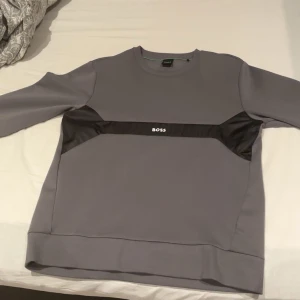Grå sweatshirt från BOSS - Säljer en stilren grå sweatshirt från BOSS med svarta detaljer över bröstet och logga framtill. Tröjan har rund halsringning och långa ärmar. Perfekt för en avslappnad och snygg look.