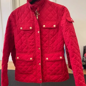 Röd quiltad jacka med guldknappar - Säljer en snygg röd quiltad jacka med guldiga knappar och fyra fickor framtill. Jackan har hög krage med spänne och markerad midja. Perfekt för dig som vill sticka ut med färg i höst eller vår.