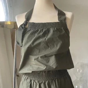 Olivgrön halterneck klänning med resår - Säljer en olivgrön klänning med halterneck och smockdetaljer vid midja och byst. Klänningen har fickor i sidorna och är tillverkad i ett lätt bomullsmaterial. Perfekt för varma dagar och enkel att styla.