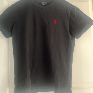 Svart t-shirt från Polo Ralph Lauren - Klassisk svart t-shirt från Polo Ralph Lauren med rund hals och korta ärmar. T-shirten har en röd broderad logga på bröstet och är tillverkad i mjuk bomull. Perfekt för en stilren och enkel look. T-shirten är mycket sparsamt använd och sitter som en storlek S. Hör av dig vid minsta funderingar