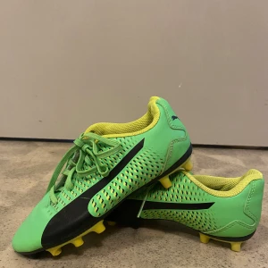 Gröna fotbollsskor från Puma - Säljer ett par gröna fotbollsskor från Puma med svarta detaljer och gula dobbar. Skorna har snörning och är designade för spel på gräs. Perfekta för dig som vill sticka ut på planen!
