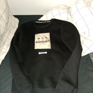 Svart sweatshirt från Burberry med beige patch - Säljer en svart sweatshirt från Burberry med en stor beige patch framtill med logga och text. Tröjan har rund halsringning och långa ärmar. Klassisk och stilren design med märkets signaturdetaljer.