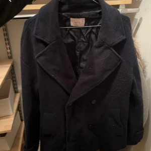 Mörkblå kappa från Nelly - Säljer en klassisk mörkblå kappa från Nelly med bred krage och knappar framtill. Jackan har två fickor och ett bälte baktill för extra detalj. Perfekt för kyligare dagar och har en stilren look.