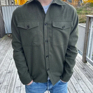 Mörkgrön overshirt från J.LINDEBERG - Stilren mörkgrön overshirt från J.LINDEBERG med klassisk krage och två bröstfickor med knappar. Skjortan har långa ärmar och är tillverkad i ett mjukt material som känns lite ulligt. Perfekt att bära som lager över en t-shirt. Nypris ligger på 2600, kom gärna med prisförslag!!