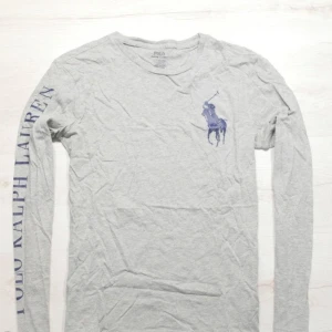 Ralph Lauren Tröja  - Säljer en ljusgrå långärmad tröja från Polo Ralph Lauren med stort blått logotryck på bröstet och text längs ena ärmen. Tröjan har rund hals och är tillverkad i mjuk bomull. Perfekt för en avslappnad och stilren look. I väldigt bra skick. Storlek S