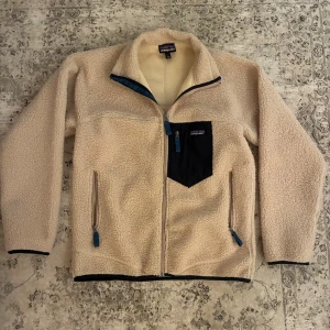 Patagonia - Mysig beige fleecejacka från Patagonia med svart bröstficka och blå detaljer på fickorna. Perfekt för kyliga dagar.