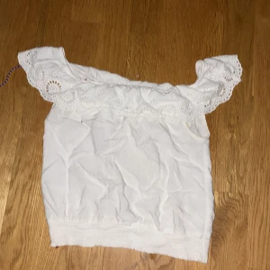 Vit offshoulder topp med broderad volang från H&M - Säljer en söt vit offshoulder topp från H&M med bred broderad volang upptill. Toppen är ärmlös och har ett romantiskt uttryck med fina hålbroderier längs volangen. Perfekt för varma dagar!