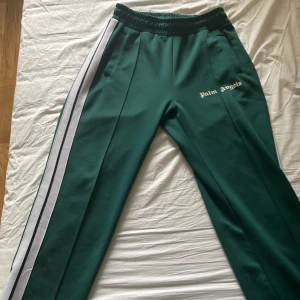 Gröna träningsbyxor från Palm Angels - Mörkgröna träningsbyxor från Palm Angels med vita sidoränder och resår i midjan. Byxorna har en sportig look och loggan broderad på vänster sida framtill. Perfekta för en avslappnad stil.