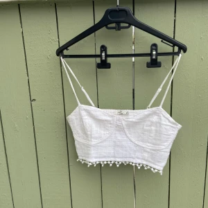 Vit croppad linnetopp från Bershka - Säljer en söt vit croppad linnetopp från Bershka med tunna axelband och dekorativa små bollar längs nederkanten. Toppen har en lätt strukturerad yta och är perfekt för varma dagar.tror den är oanvänd.