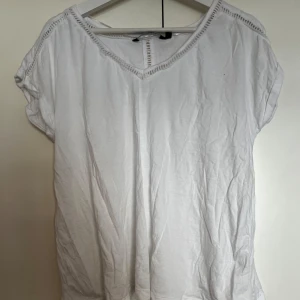 Vit blus med hål detaljer - Säljer en vit blus med korta ärmar och v-ringning. Blusen har fina  detaljer längs halsringningen och axlarna som ger en extra touch. Perfekt till sommaren och enkel att matcha med olika outfits.