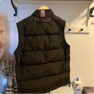 Svart dunväst från Parajumpers - Säljer en svart dunväst från Parajumpers med hög krage och dragkedja framtill. Västen har två stora fickor med knappar och dragkedjor samt en gul detalj vid halsen. Perfekt för lager-på-lager under kyliga dagar.