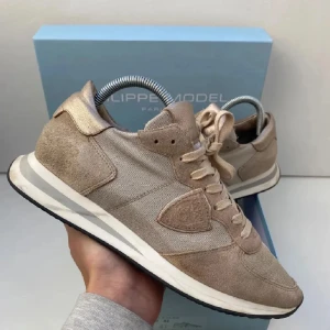 Beige sneakers från Philippe Model - Säljer ett par snygga beige sneakers från Philippe Model. Skorna har en mix av mocka och textil, med vita detaljer på sulan och klassisk snörning. Perfekta för dig som gillar stilrena och neutrala färger.