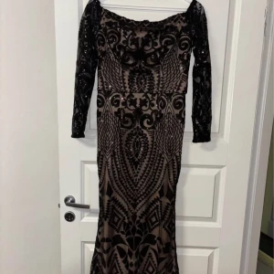 Svart spetsklänning i off-shouldermodell från Dennis Maglic - Elegant svart långklänning från Dennis Maglic med långa ärmar och vackert mönster av paljetter. Klänningen har ”transparenta” partier och figurnära passform. Använd en gång 