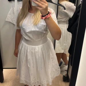 Vit klänning med broderade detaljer - Säljer en söt vit klänning med korta ärmar och broderade hålbroderier längs ärmar och fåll. Klänningen har en markerad midja med resår och en luftig kjol. Perfekt för sommarens alla tillfällen! Oanvänd med prislapp kvar!💕