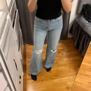 HELT NYA! Blå vida jeans med slitningar lågmidjade - Ett par ljusblå jeans från H&M med vida ben och slitningar på båda knäna. Jeansen har klassisk femficksdesign och knappar i gylfen. Helt nya och oanvända med alla prislappar kvar. Storlek 34 och heter Low waist Wide full length.