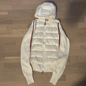 Vit Moncler cardigan - Säljer en vit cardigan från Moncler med huva. Jackan har stickade ärmar med röd och svart rand längs sidorna. Storlek L men passar M