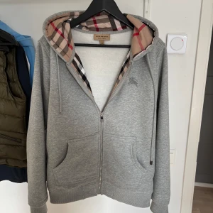 Grå zip-hoodie från Burberry - Grå hoodie med dragkedja från Burberry. Klassisk rutigt foder i huvan och två fickor framtill. Mjukt material och justerbar huva med snören. Perfekt för en stilren och avslappnad look.