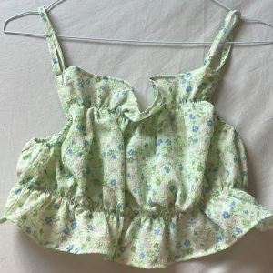 Ljusgrön blommig croppad linnetopp - Superfin ljusgrön croppad linnetopp med blommigt mönster i blått och rosa. Toppen har smala axelband och volangdetaljer nedtill. Perfekt för varma dagar och ger en söt och somrig look.