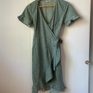 Grön omlottklänning med blommigt mönster från ONLY - Säljer en ljusgrön omlottklänning från ONLY med små mörkgröna blommor. Klänningen har korta ärmar, volangdetalj nertill och knyt i midjan. Perfekt för vår och sommar!