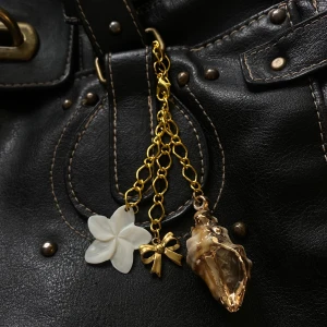 Guldfärgad bagcharm med snäcka, blomma och rosett - Unik bagcharm med grov guldfärgad kedja och tre hängen: en vit blomma, en liten rosett och en snäcka i guldton. Perfekt för dig som gillar detaljer och strandinspirerad stil.