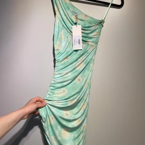 Monza Dress från Adoore - Säljer en ljusgrön Monza-klänning från Adoore med blommigt mönster. Klänningen har oneshoulder-design och går ner till anklarna. Aldrig använd