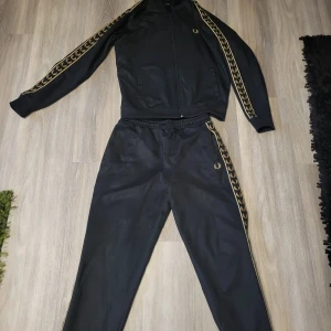 Svart tracksuit från Fred Perry med gulddetaljer - Säljer tracksuit från Fred Perry med guldfärgade loggor längs sidorna. Byxorna har resår i midjan och en smal passform. Perfekta för en sportig look.