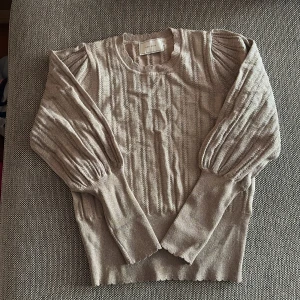 Beige stickad tröja med puffärm från Neo Noir - Supersnygg beige stickad tröja från Neo Noir med puffärmar och diskret mönsterstickning framtill. Glitter tyg. Tröjan har rund halsringning och ribbade muddar vid ärmslut och nederkant. Perfekt för lager-på-lager och stilrena outfits. Använt vid ett tillfälle.