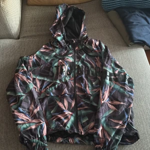 Mönstrad vindjacka med huva från Stronger - Säljer en snygg vindjacka från Stronger i storlek medium. Jackan har ett tropiskt bladmönster i grönt, rosa och svart, dragkedja framtill, huva och resår vid ärmslut och nederkant. Perfekt för dig som vill tex springa ute.