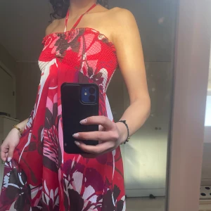 Röd och vit maxiklänning med blommigt mönster - Säljer en långklänning med halterneck och blommigt mönster i rött, vitt och svart. Klänningen har en smockad överdel och faller luftigt ner till golvet. Perfekt för sommardagar!Faller över hälen på mig som är 172💃