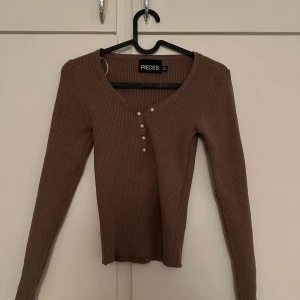 Beige ribbad topp med knappar från Pieces - Säljer en beige långärmad topp från Pieces med ribbad struktur och dekorativa knappar framtill. Toppen har en djupare rund halsringning och är perfekt för lager-på-lager-stil.