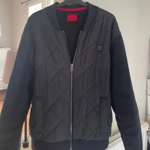 Svart skön cardigan från Hugo Boss - Säljer en svart cardigan från Hugo Boss med quiltat mönster framtill och släta armar. Står strl M men passar även strl L. Perfekt för dig som gillar stilren och modern design.