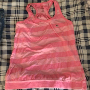  linnetopp från Lacoste - Säljer en rosa och ljusrosa randig linnetopp från Lacoste med klassisk broderad krokodillogga på bröstet. Modellen är ärmlös med brottarrygg och har en sportig känsla. Perfekt för varma dagar eller träning.