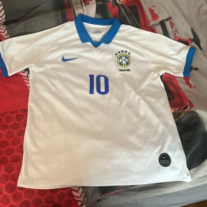 Vit Brasilien fotbollströja Neymar JR 10 - Snygg vit Brasilien fotbollströja med blå detaljer och Neymar JR 10 på ryggen. Tröjan har blå krage, blå Nike-logga och Brasilien-emblem på bröstet. Perfekt för dig som vill visa ditt stöd för landslaget. Storlek L men passar även de som har M